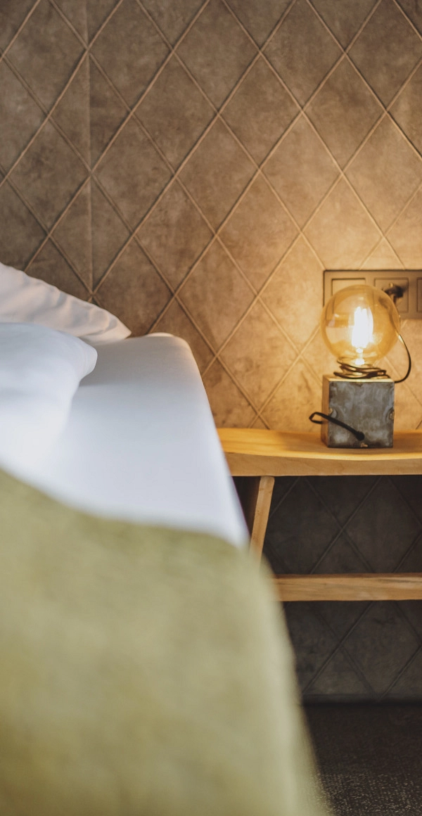 Bett mit Kopfkissen und Lampe auf einem Holztisch, sowie einem strukturierten Wandhintergrund.